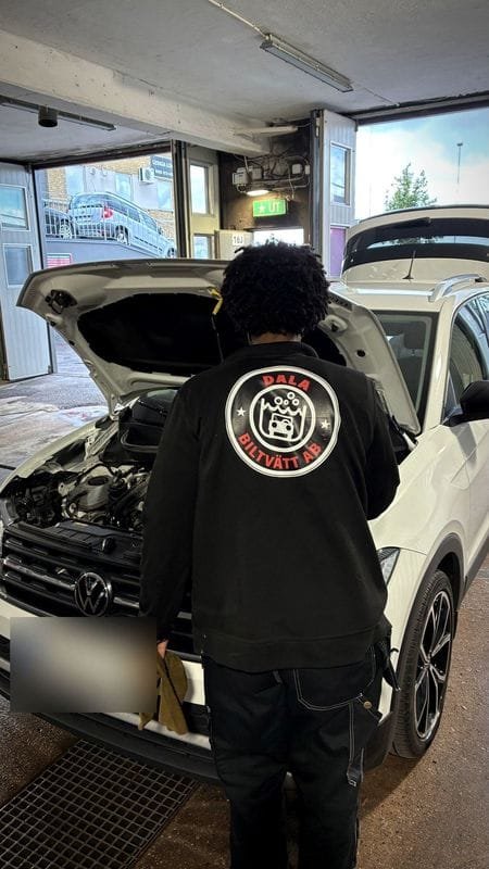 En medarbetare, med Dala Biltvätts logotyp på ryggen, framför en vit bil med öppen motorhuv.