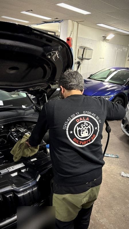 En medarbetare i Dala Biltvätt-tröja lutar sig in över motorrummet på en bil med öppen motorhuv.